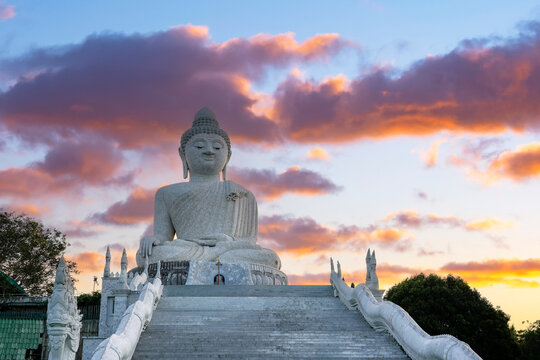 The colossal white marble Big Buddha statue sits calmly on Nakkerd Hill, Phuket, Thailand. Vibrant orange and purple sunset clouds drift across the sky.