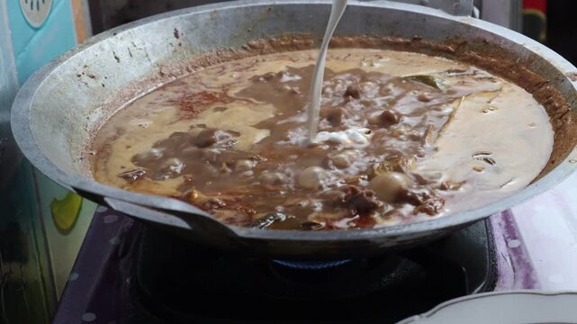 Thick white coconut milk being poured into a large traditional wok filled with simmering Indonesian beef rendang and aromatic spices.