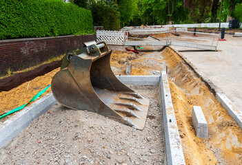 Large excavator bucket rests on gravel at a road construction site, with sand trenches, concrete curbs, temporary barriers, brick wall, and urban infrastructure work. © Volha