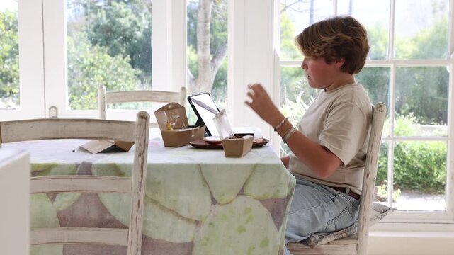 12 year old boy eating box lunch in kitchen using tablet computer