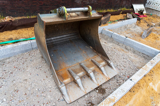 Close view of a heavy excavator bucket with metal teeth on a gravel roadwork area, showing scratched steel, rust marks, sand trench, concrete curbs, and construction detail.