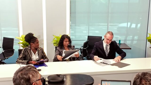 Diverse professionals meet in a modern conference room, reviewing documents and tablets at a long table with sleek chairs, glass walls, and plants during a collaborative discussion.