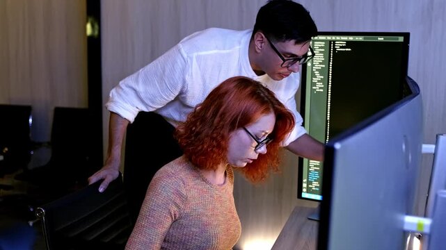 Two colleagues collaborate at a workstation, reviewing code on dual monitors in a modern office, focused on debugging and software development