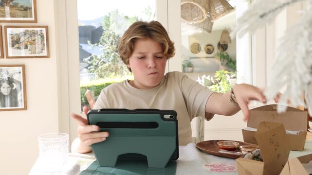 12 year old boy eating box lunch in kitchen using tablet computer