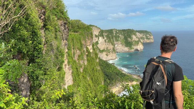 A young male traveler standing on a cliff edge overlooking the dramatic coastline of Nusa Penida Island, Bali. Back view, no face visible. Turquoise ocean, limestone cliffs, and green hills. Slow moti