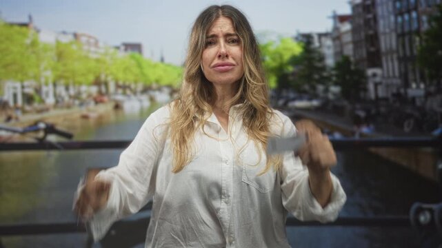 Woman holding a creditcard in a raised hand while hand on head gesture on a street by a canal and bicycle; financial worry frustration.