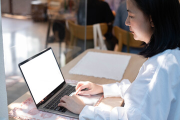 Fototapeta premium Asian woman typing on laptop with blank screen in cafe, side view, remote work lifestyle, digital workspace mockup, modern business, productivity and online communication with soft natural light.