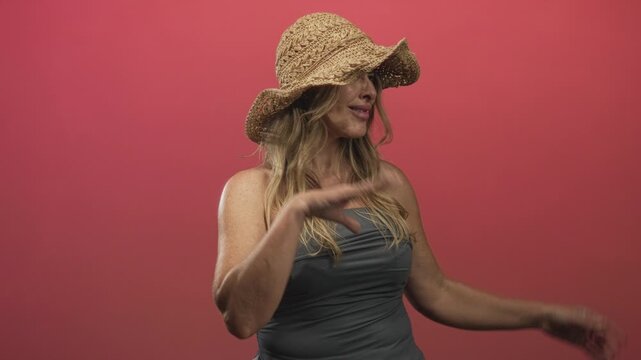 Woman presents open hands and smiles wearing a straw hat and strapless top in studio; playful confidence.
