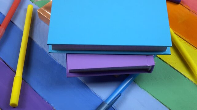 Multicolored notepads and ballpoint pens lie on a wooden rainbow colors board, top view  