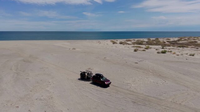 Watch as an SUV pulls a teardrop camper across sandy roads in Baja California, Mexico under a blue sky by the coast.