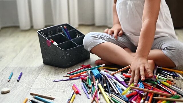 Child collecting colorful pencil from wooden floor near storage box, small hands gathering scattered pencil after drawing activity. crayons cleanup during creative play, childhood art, concept