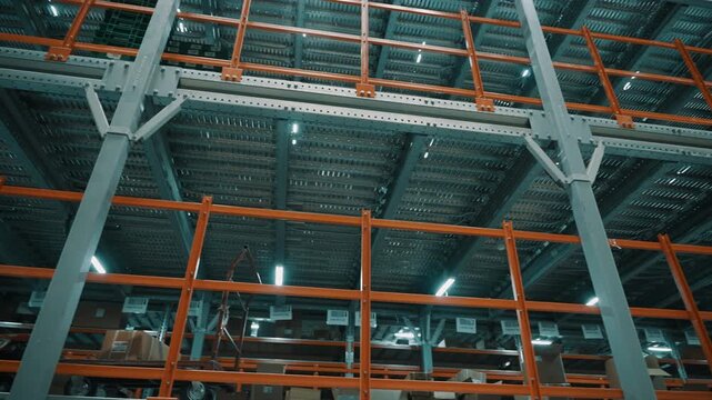 Industrial warehouse interior with multi-tier steel pallet racking and orange shelving, illuminated by overhead lighting beneath ceiling trusses, beams, and supports