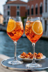 Naklejka premium A realistic commercial stock photo of two bright orange Aperol Spritz glasses on a small round metal cafe table, Venetian canal and historic architecture in the background, late af