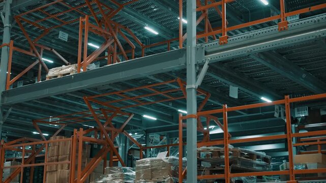 Multi-level warehouse racking with orange safety rails, stacked pallets, and cardboard boxes under bright LED lighting in a modern logistics and distribution facility.