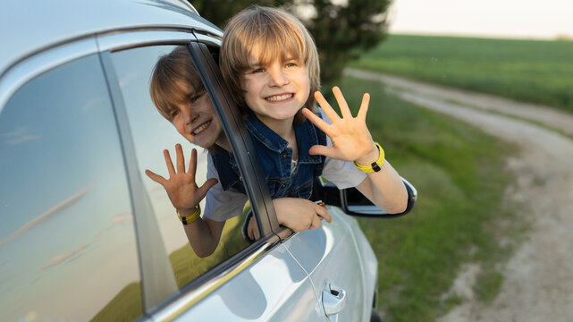 pre-teen boy looks out of the car window and waves his hand cheerfully, enjoying the fresh air and a road trip on vacation. The child's face is reflected in the window.