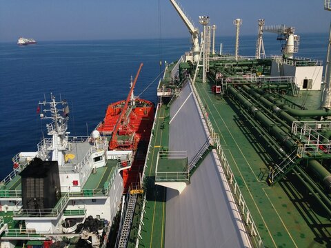 A large liquefied gas carrier deck connects to a smaller red bunkering vessel on the open sea. The perspective highlights industrial pipelines, ship structures, and a clear blue ocean horizon.