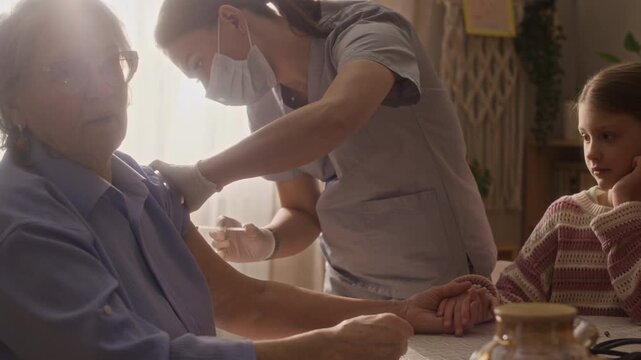 Medium shot of paramedic giving injection to elderly woman who holding granddaughters hand