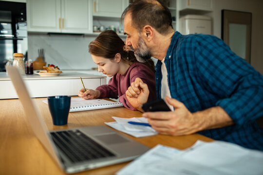 Parent multitasking with bills while child does homework at kitchen table