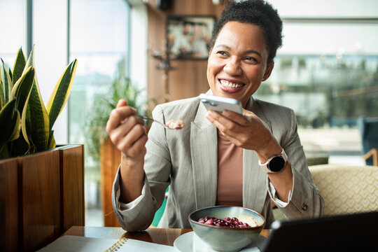 Smiling businesswoman eating breakfast and using smartphone in cafe