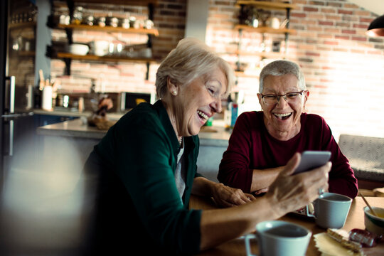 Two senior women laughing at smartphone in home kitchen