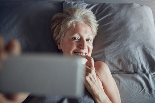 Smiling senior woman taking selfie in bed at home
