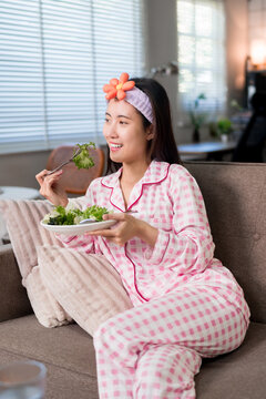 healthy Asian women eating fresh salad vegetable for breakfast morning routine at home sitting on sofa wearing pink pajamas relaxing wellness lifestyle diet