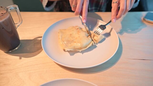 A person using a fork and knife cuts a homemade dish on a plate at a dinner table. The focus is on the activity of enjoying food with casual dining elements present