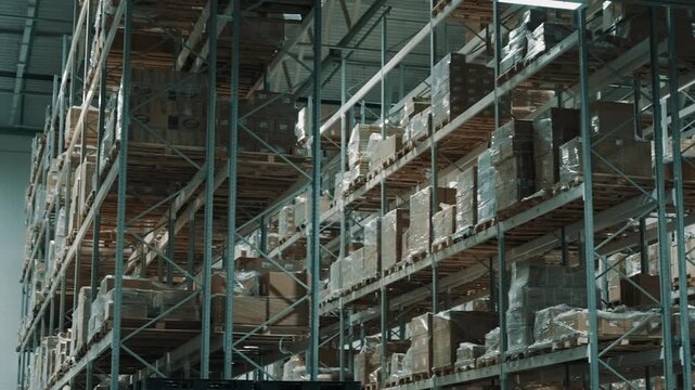 Industrial warehouse interior with high steel racking stacked with palletized cardboard boxes and shrink-wrapped goods, organized inventory under bright overhead lights