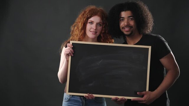 A diverse couple holds a blank chalkboard, offering ample copy space for a customizable message. The happy and friendly expressions of the man and woman convey a positive mood, making it various comme