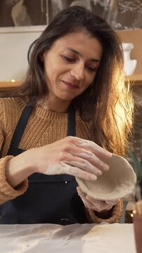 Caucasian woman carefully shapes the rim of a clay mug with her fingers in a cozy workshop. The process of handcrafting, ceramics creation, hobby, and leisure concepts.