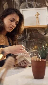 A Caucasian woman carefully shapes the rim of a clay mug with her fingers in a cozy workshop. The process of handcrafting, ceramics creation, hobby, and leisure concepts