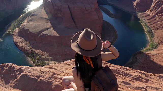 Cheerful young woman explorer sitting on the cliff edge, smiling at the camera and admiring the stunning horseshoe bend of the colorado river near page, arizona on a sunny summer day