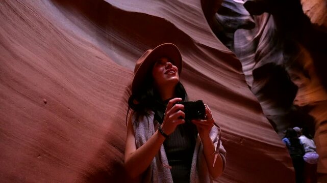 Young smiling woman with a camera wearing a fedora hat looking up and photographing the stunning red rock formations of a narrow slot canyon in arizona during her american vacation
