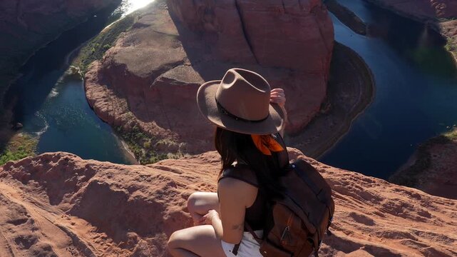 Smiling woman with a backpack sitting on the edge of a cliff, looking at the camera and then raising her arms in freedom while observing the majestic horseshoe bend in page, arizona
