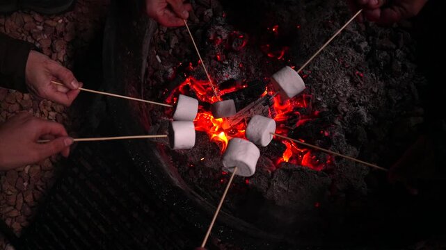Group of friends roasting sweet and tasty marshmallows on wooden skewers over a bonfire at night, enjoying a warm and cozy evening around the glowing embers of a campfire in the darkness
