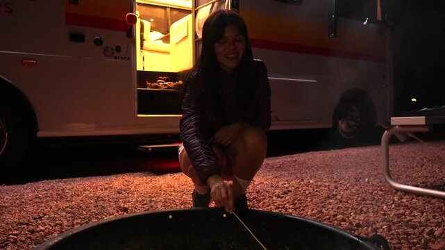 Young woman enjoying a camping trip at night, crouching by a fire pit and roasting a marshmallow on a stick over glowing embers with her camper van illuminated in the background