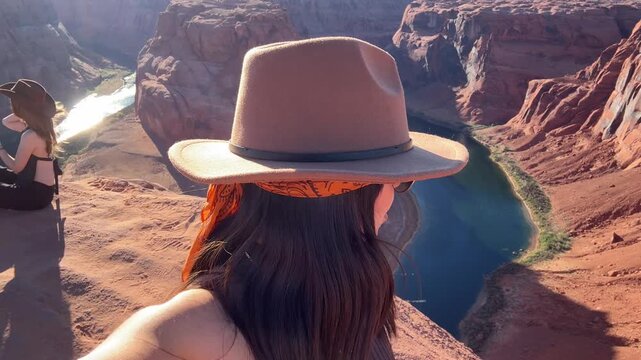 Stylish young woman wearing a hat and sunglasses filming herself with a smartphone, showing the breathtaking panorama of horseshoe bend on the colorado river in page, arizona