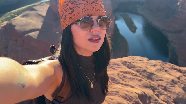 Stylish young woman with sunglasses and a bandana taking a selfie video at the stunning horseshoe bend overlook, showing the breathtaking canyon and colorado river landscape in page, arizona