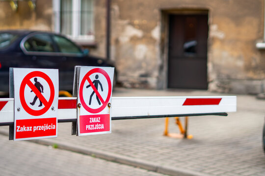 White and red automatic barrier gate arm with two no pedestrian entry signs and Polish text Zakaz przejścia, captured in a close-up shot with a blurred courtyard background.