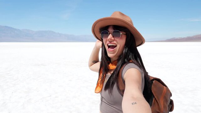 Young smiling woman with a hat taking a selfie in a vast, white salt flat on a sunny day