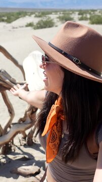 Happy young female explorer with a hat and sunglasses enjoying a sunny day in the mesquite flat sand dunes of death valley national park, california, smiling and looking at the camera