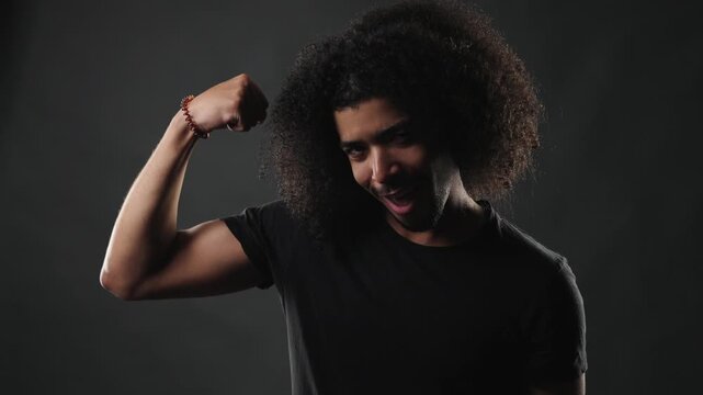 A serious young African American man with voluminous curly hair and a neatly trimmed beard is captured in a studio portrait. He wears a simple black t shirt, which contrasts subtly with the dark backg