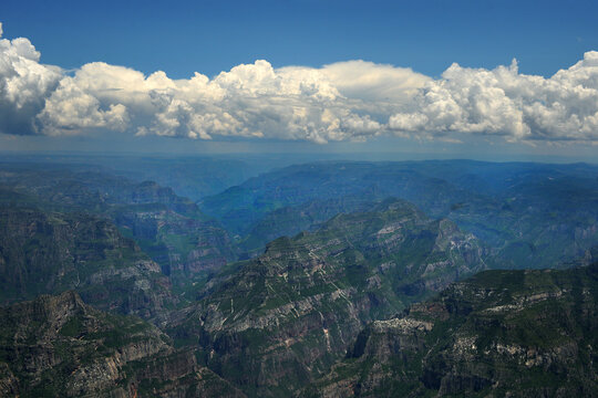 Aerial view of Barrancas del Cobre featuring vast canyon systems with green slopes and rocky cliffs under white clouds in Chihuahua, Chihuahua, Mexico.