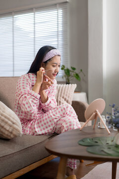 Asian woman applying face mask skincare beauty treatment morning routine sitting on sofa at home wearing pink pajamas relaxing in living room weekend wellness
