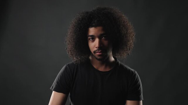 A young adult man with a voluminous afro hairstyle and a dark t shirt poses confidently against a dark, plain background. His serious expression and direct gaze into the camera convey a sense of self 