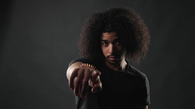 A serious young man with a voluminous afro hairstyle and a dark t shirt looks directly at the camera with an intense and thoughtful expression. The studio portrait features dramatic lighting, casting 