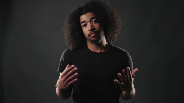 A serious young adult man with voluminous curly hair and a plain black t shirt stands against a dark, minimalist background. He looks directly at the viewer with a neutral and thoughtful expression, c