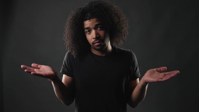 A confident young man with voluminous curly hair and a plain black t shirt stands against a dark, moody background. He looks directly at the camera with a serious and composed expression, conveying a 