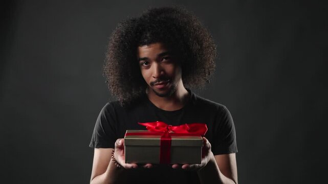 A thoughtful gift giving concept. A young man with curly hair holds out a small present box adorned with a red ribbon against a dark background, conveying a sense of generosity and care. This footage 