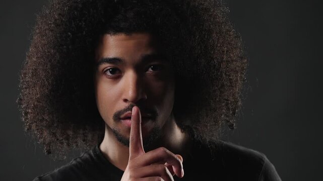 This depicts a concept of secrecy and confidentiality, a young man with an afro hairstyle gesturing for silence. He places his finger on his lips in a close up studio shot against a dark background.
